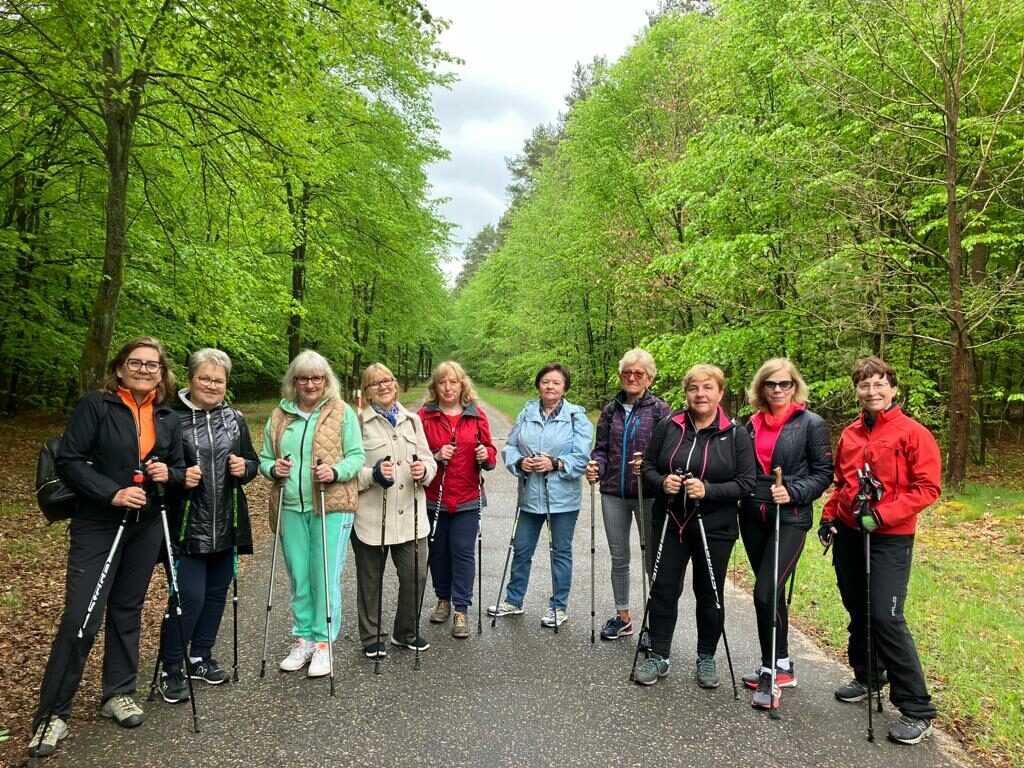 Na zdjęciu widać: Grupa Aktywni 55 plus. Kobiety z kijkami trekkingowymi. Z tyłu widać aleję zielonych drzew.