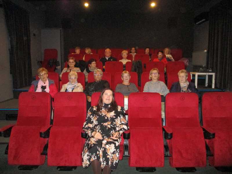 Seniorzy w kameralnej sali kinowej. Po trzy fotele w rzędzie, a rzędów widać pięć. Fotele w czerwonym kolorze. 